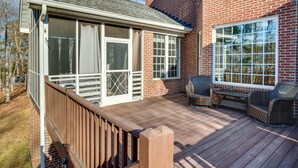 A covered deck with a wooden railing, a brick building with large windows, and wicker furniture. - Executive Wfh-friendly Haven in Blythewood! (Blythewood)