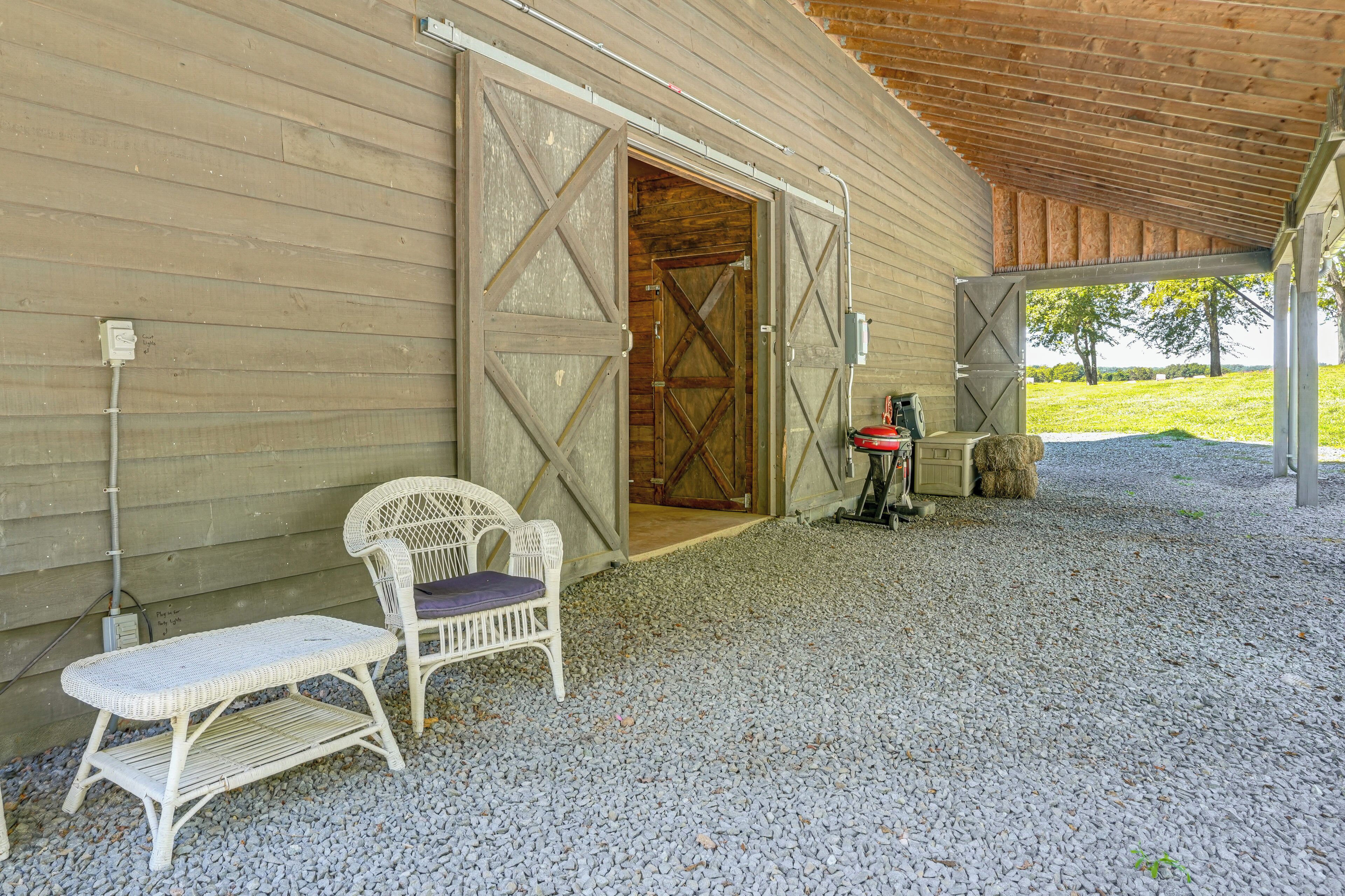 A covered outdoor seating area with a wicker chair and table, a barn-style door, and a gravel ground.