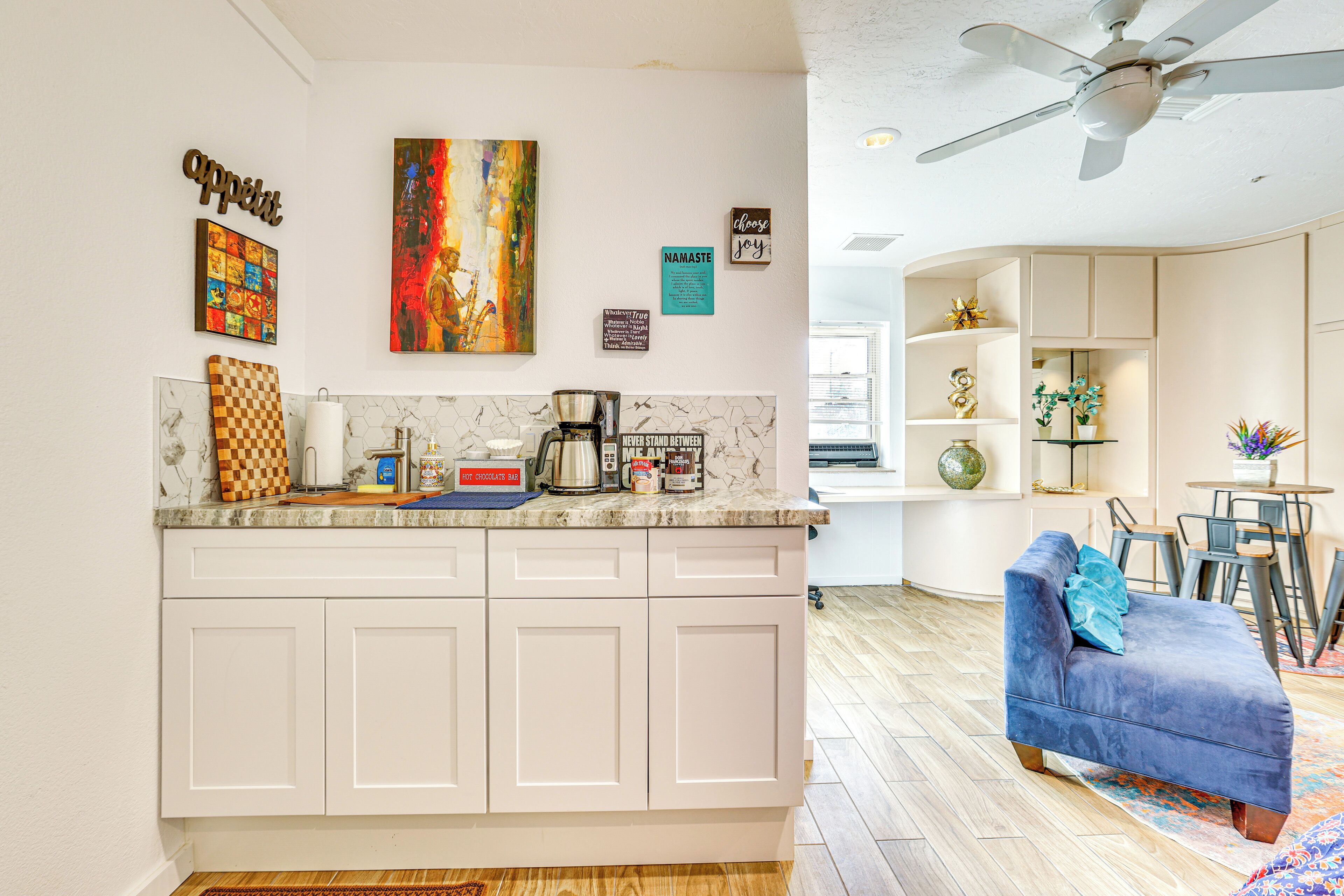 A modern kitchen with white cabinets, a coffee maker, and a checkered board. There is a blue sofa in the living area with a decorative pillow.