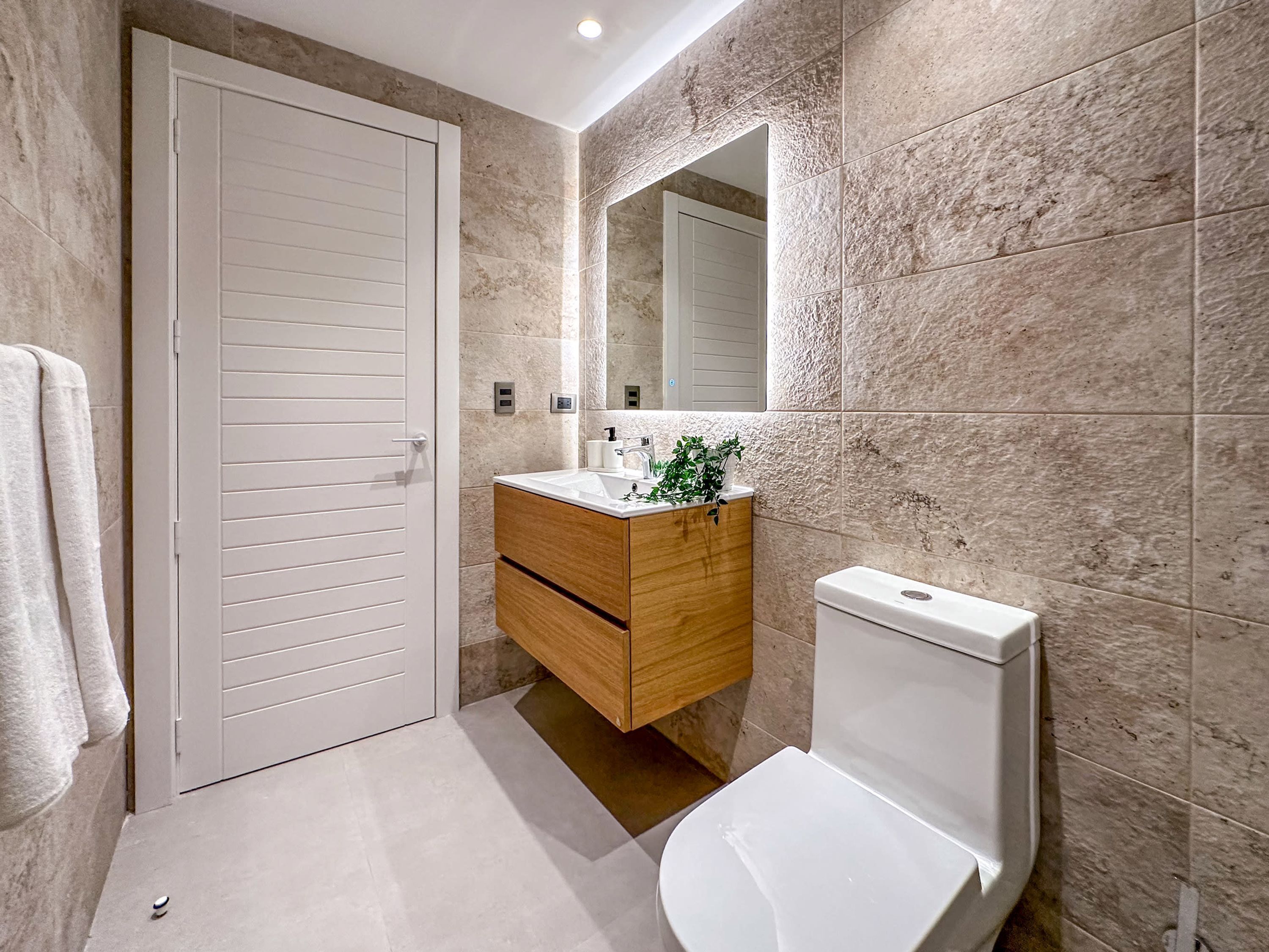 A bathroom with a white toilet, a wooden vanity, a mirror, and beige tiled walls.