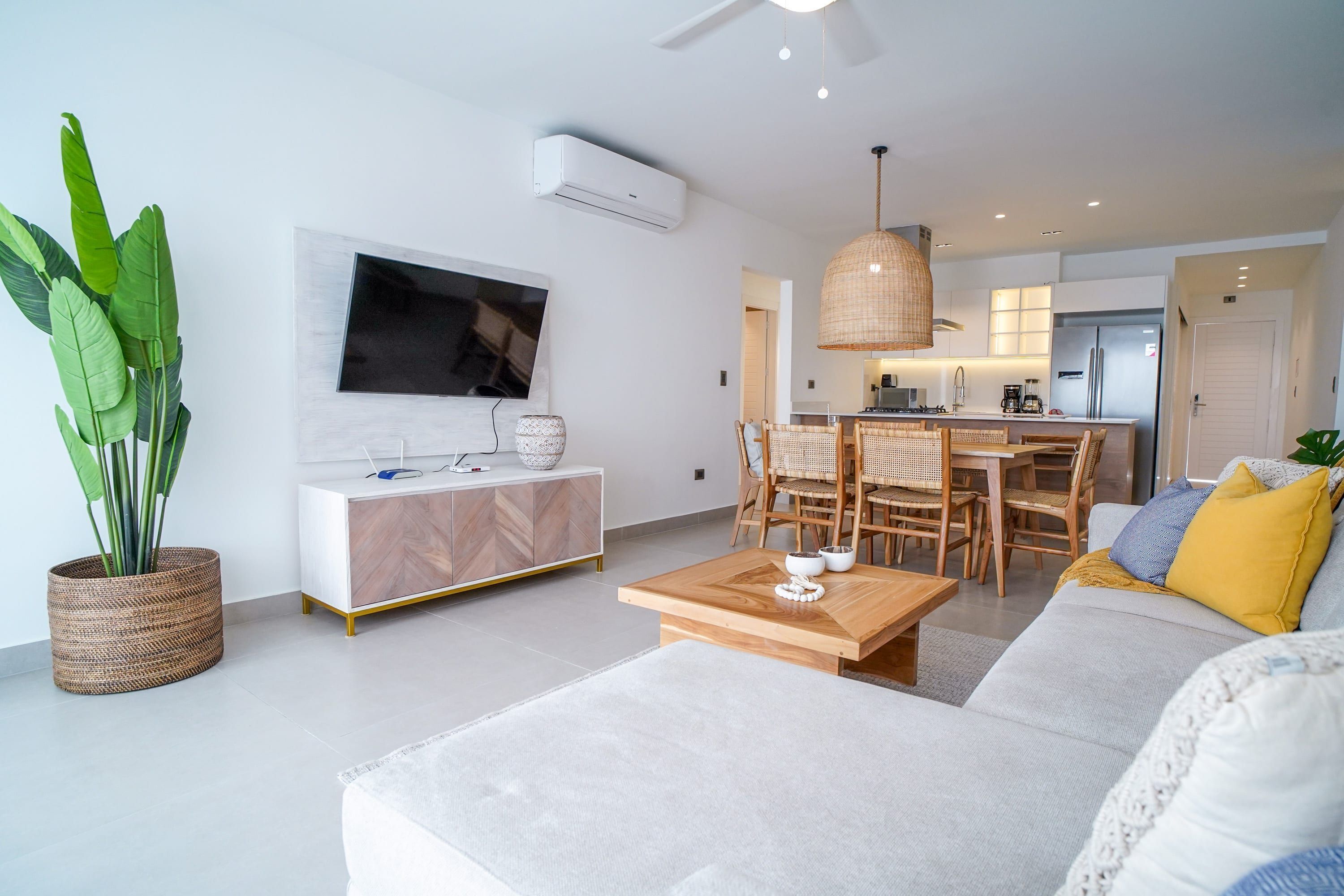 A modern living room with a sofa, coffee table, and a TV mounted on the wall. There is a dining area with a table and chairs, and a kitchen in the background.