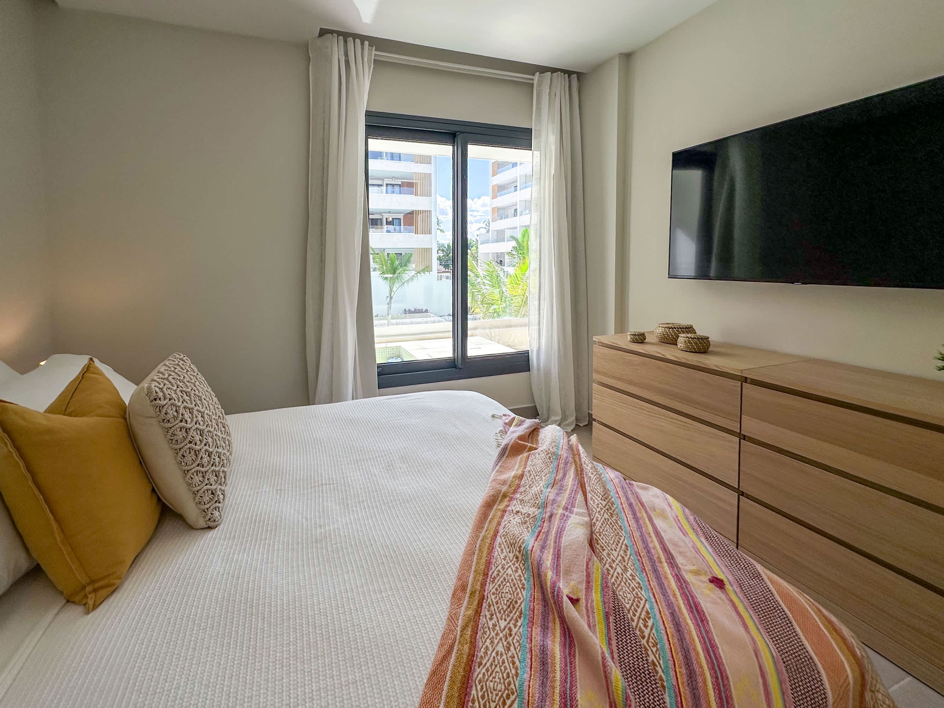Een hotelkamer met een bed, een televisie, een dressoir en een raam met uitzicht op gebouwen en groen.