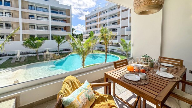 A balcony with a wooden table set for breakfast, overlooking a swimming pool and modern buildings. - Spectacular Brand new Beach Condo (Punta Cana)