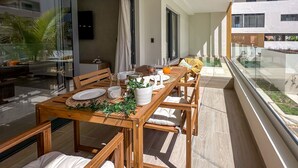 A modern outdoor dining area with a wooden table and chairs, a glass railing, and a view of a swimming pool and buildings. - Spectacular Brand new Beach Condo (Punta Cana)