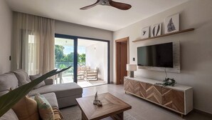 A modern living room with a sofa, coffee table, TV, and a balcony with outdoor furniture. - Spectacular Golf Front 3BR Apartment! (Punta Cana)