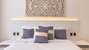 Une chambre moderne avec une tête de lit blanche, un panneau décoratif mural et un lit recouvert d’une literie blanche agrémentée d’oreillers à motifs.