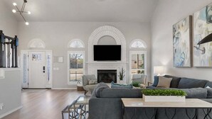 A spacious living room with a fireplace, a large flat-screen TV mounted above it, a grey sofa, and a coffee table with a plant. - South Tulsa Splash W/Pool & Hot Tub (Tulsa)