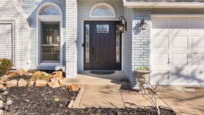 A house with a dark front door, two windows with arched tops, a garage door, and a small garden area with rocks and a potted plant. - South Tulsa Splash W/Pool & Hot Tub (Tulsa)