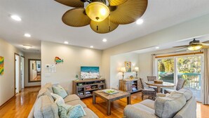 A spacious living room with a wooden floor, a ceiling fan, a television, and a dining area with a table and chairs. - Sassy Seahorse: Heated pool, hot tub, ping pong, Foosball and pool table! (Anna Maria)