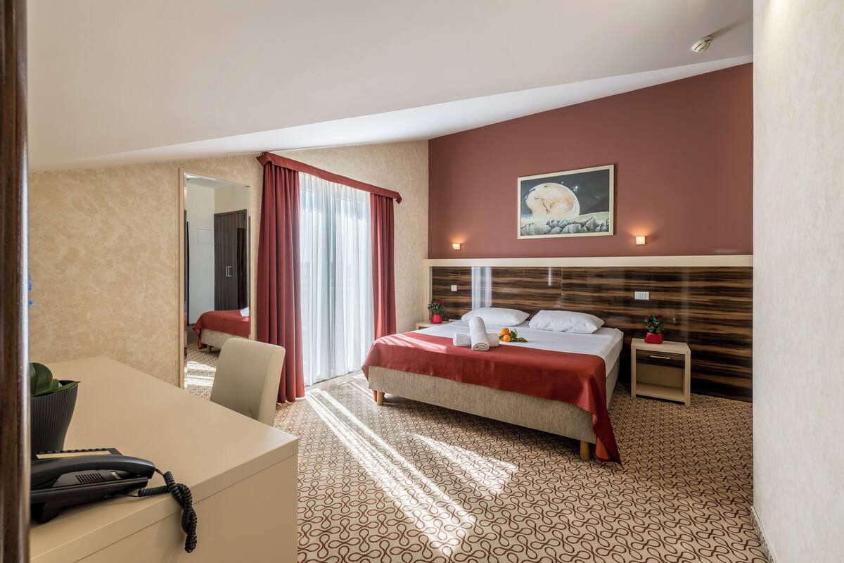 A hotel room with a bed, a desk with a telephone, a nightstand with a lamp, and a painting on the wall.