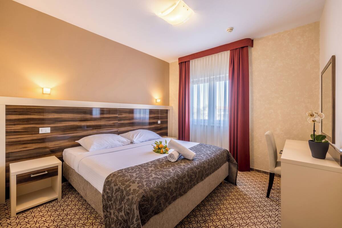 A hotel room with a bed, bedside tables, a desk, a chair, and a potted plant.