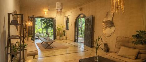 Amplia sala de estar con sofá, mesa de centro y plantas en macetas. Se observa una escalera de madera y una lámpara colgante.