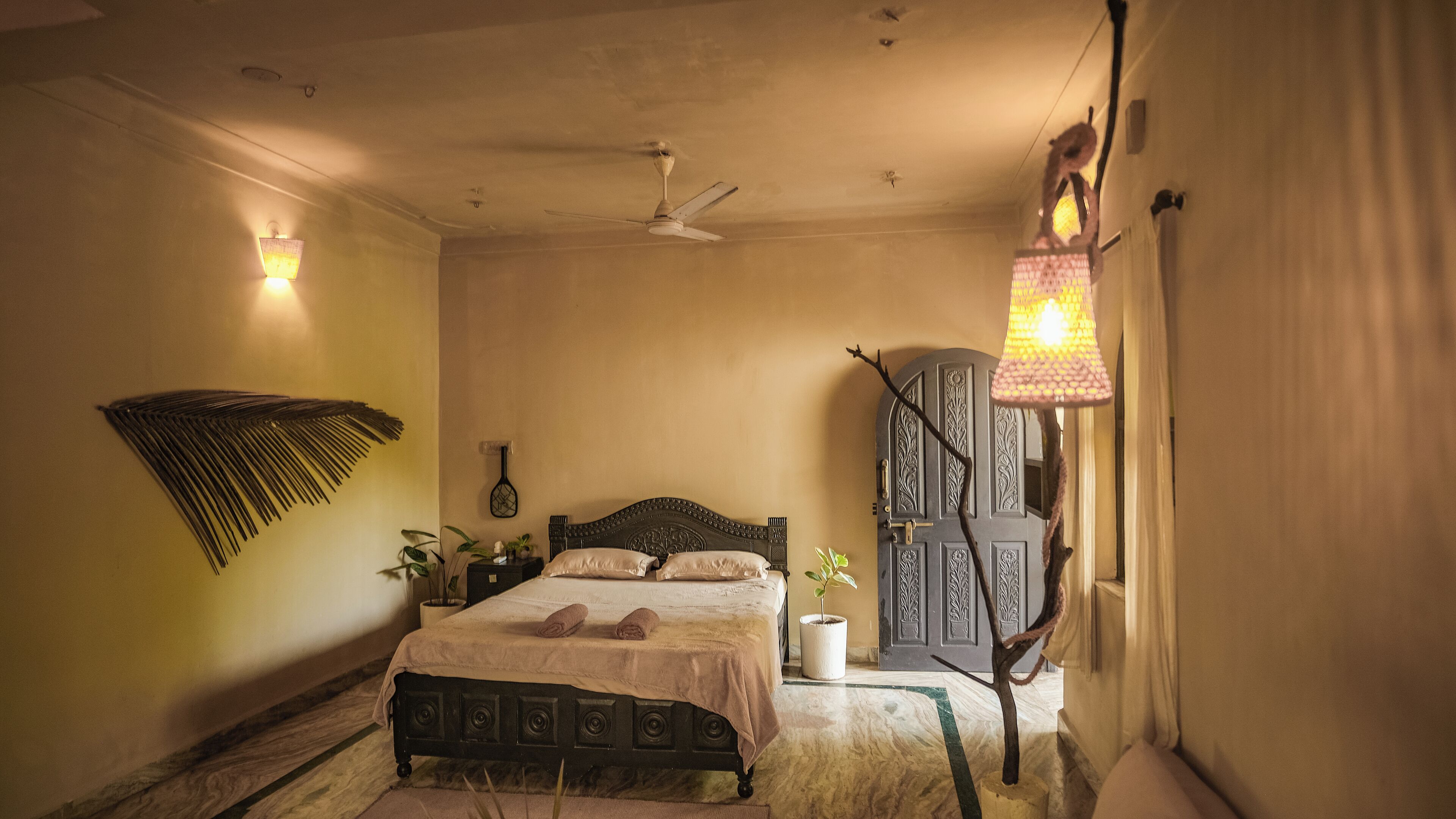 A bedroom with a bed, a ceiling fan, a wall-mounted lamp, and a decorative plant.