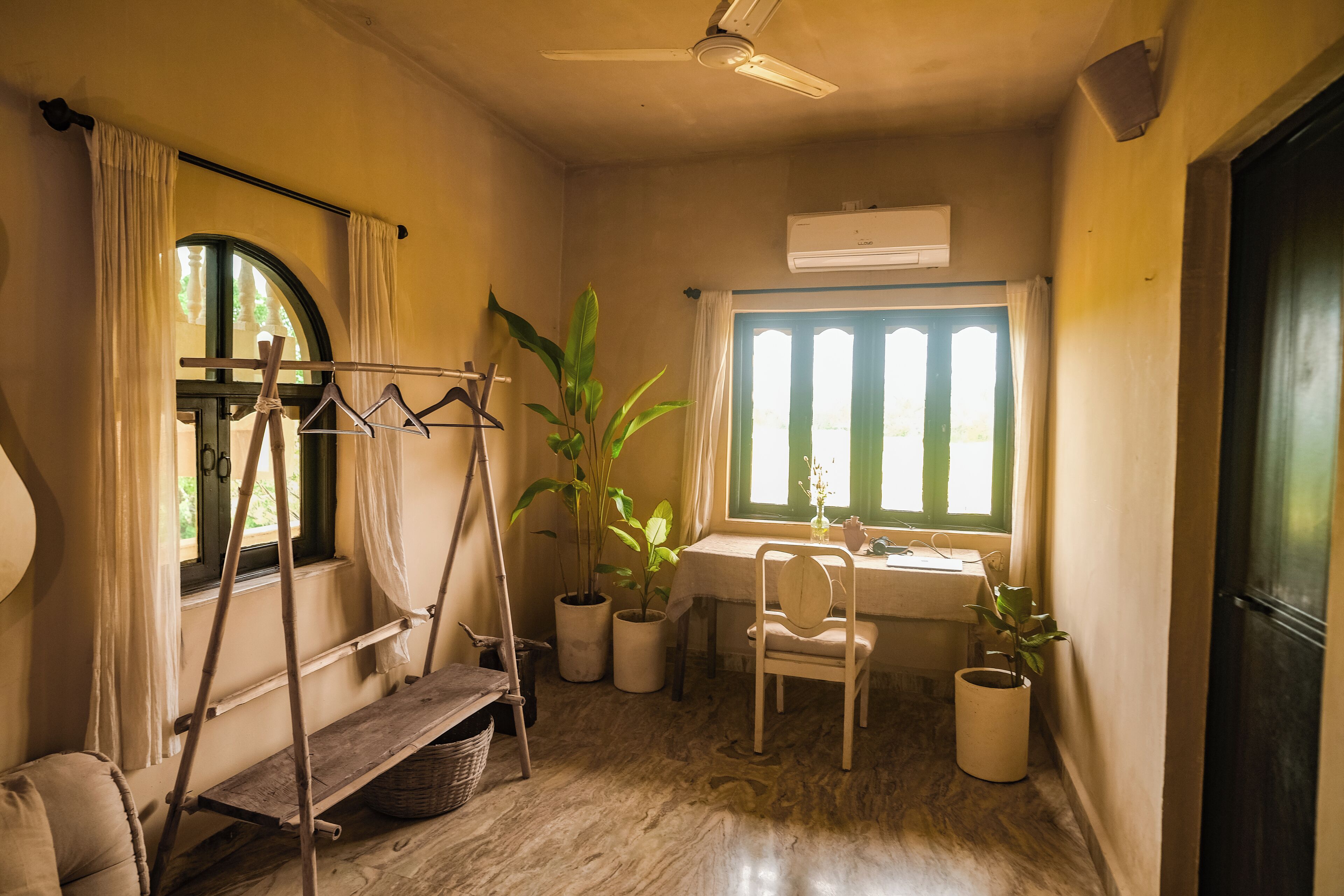 Une pièce avec un plancher en bois, un ventilateur de plafond, une fenêtre avec des rideaux, un bureau avec une chaise et plusieurs plantes en pot.
