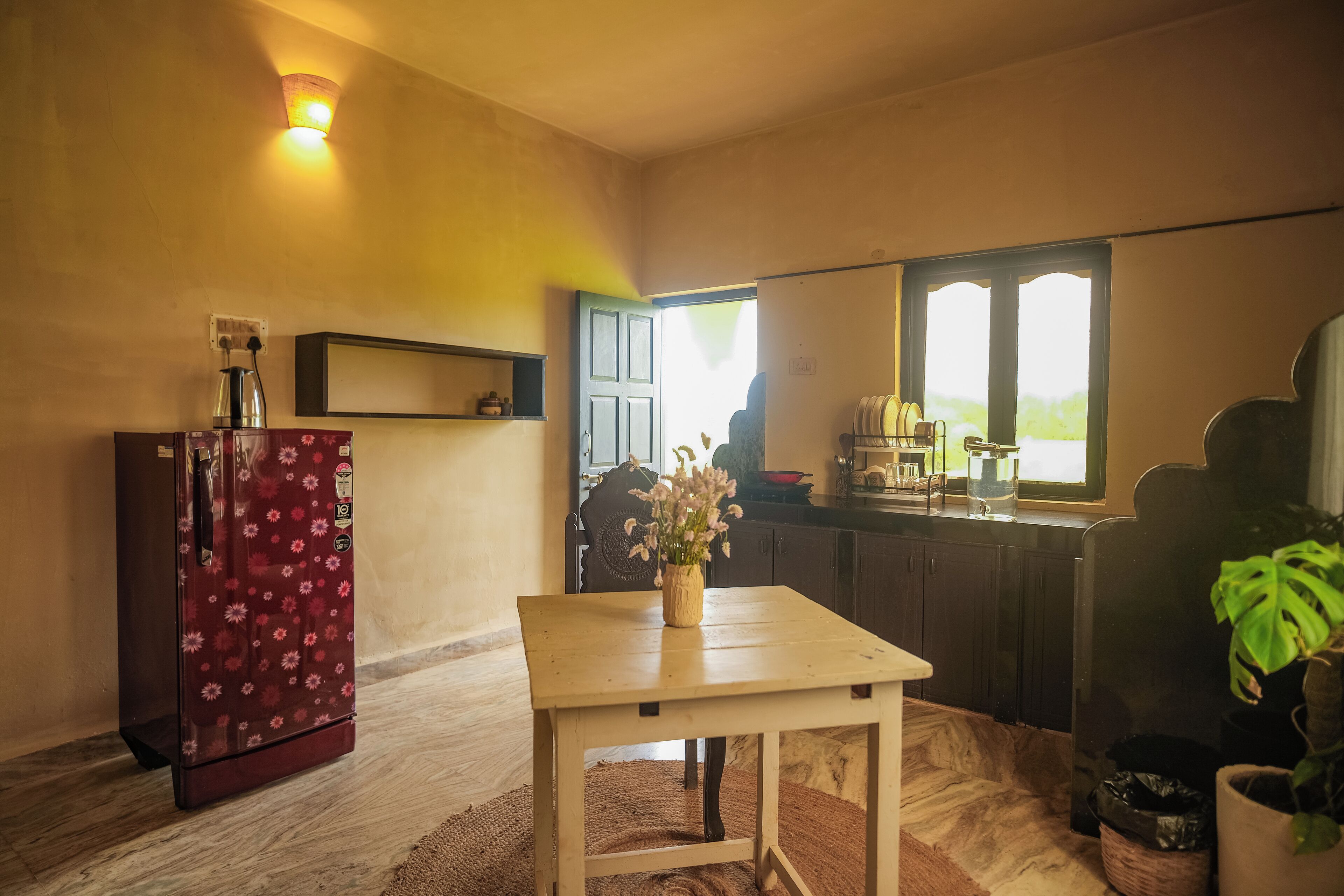Una cocina compacta con refrigerador, una mesa de madera con un florero, una ventana y una puerta que da a otra habitación.