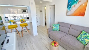 A modern living room with a grey sofa and tropical patterned cushions, a dining area with yellow chairs, and a kitchen with white cabinets and a microwave. - Vacation apartment Costa Sun, Costa Calma, pool, WiFi, balcony (Costa Calma)
