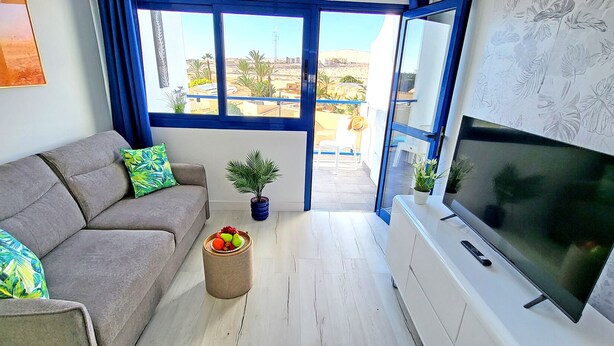 A living room with a grey sofa, a flat-screen TV, and a balcony with a view of buildings and palm trees. - Vacation apartment Costa Sun, Costa Calma, pool, WiFi, balcony (Costa Calma)
