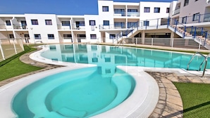 A swimming pool with a curved design, surrounded by a well-maintained lawn and a building with multiple balconies and stairs. - Vacation apartment Costa Sun, Costa Calma, pool, WiFi, balcony (Costa Calma)