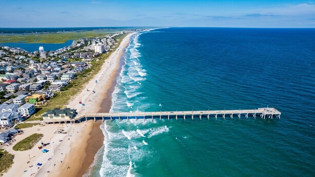 Vista aérea de una playa con un muelle largo que se adentra en el océano, rodeada de edificios residenciales y una orilla arenosa.