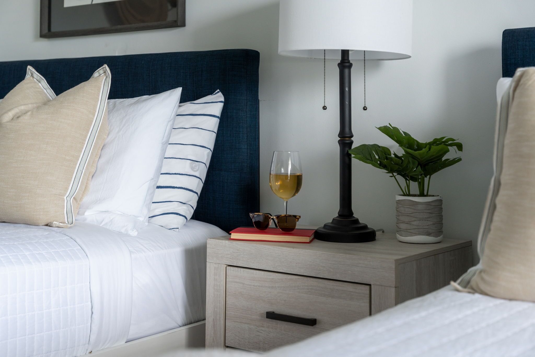 Una cama bien hecha con ropa blanca y almohadas a rayas, una mesita de noche con una lámpara, un libro, una copa de vino y una planta en maceta.