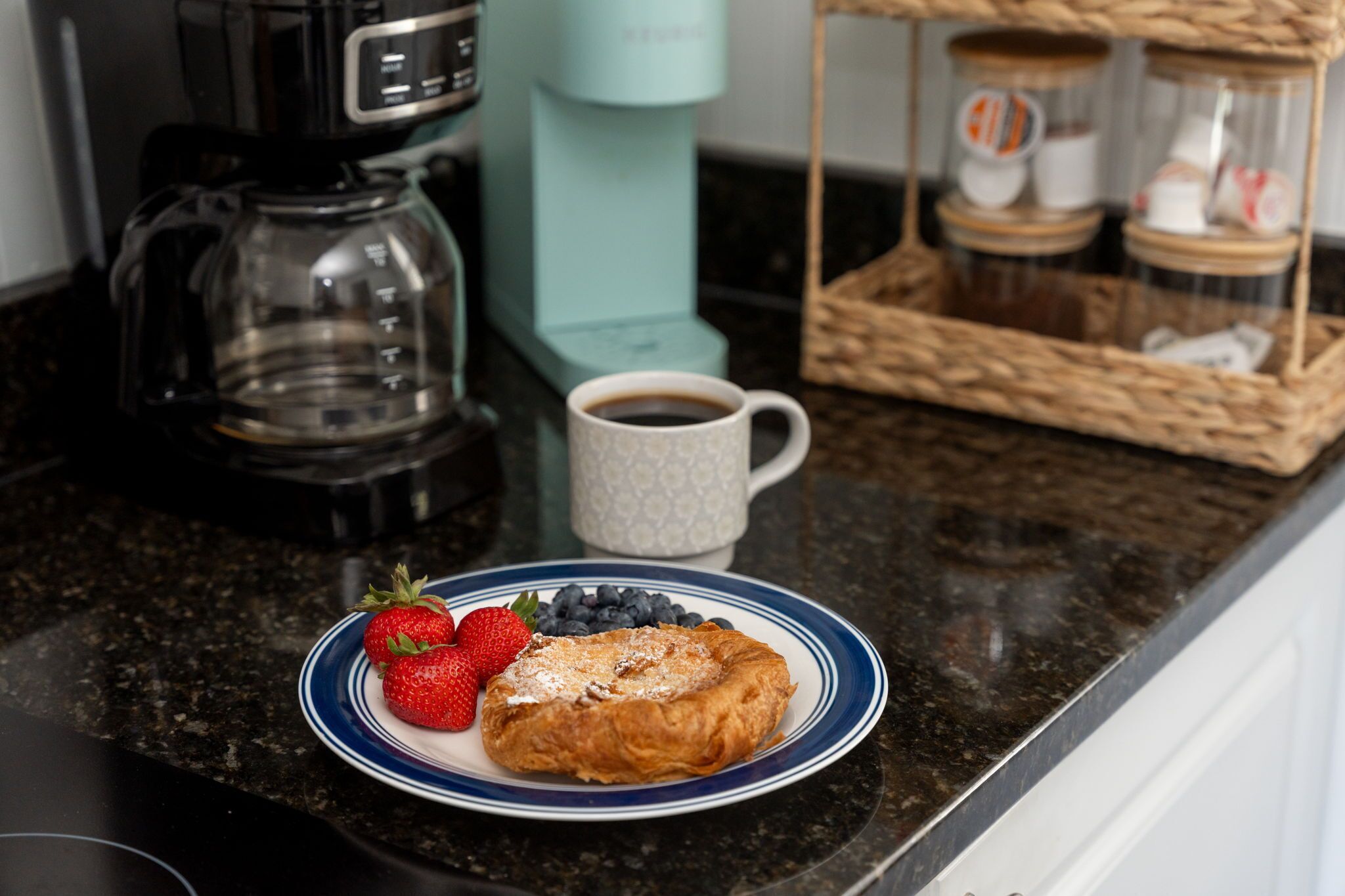 Une table mise pour le petit-déjeuner, avec une assiette de nourriture, une machine à café et un panier rempli de pots.