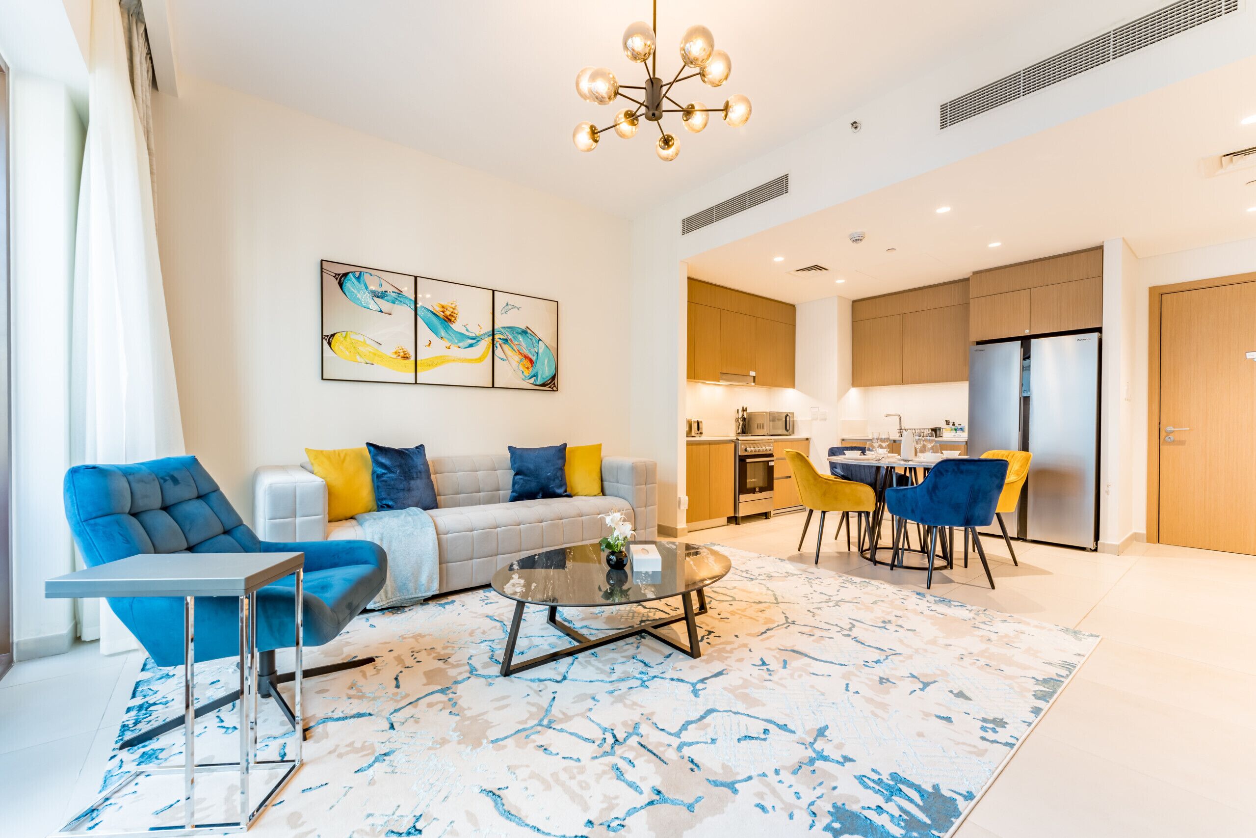 Una sala moderna con sofá, mesa de centro y sillas, un área de cocina con armarios y refrigerador, y una mesa de comedor con sillas.