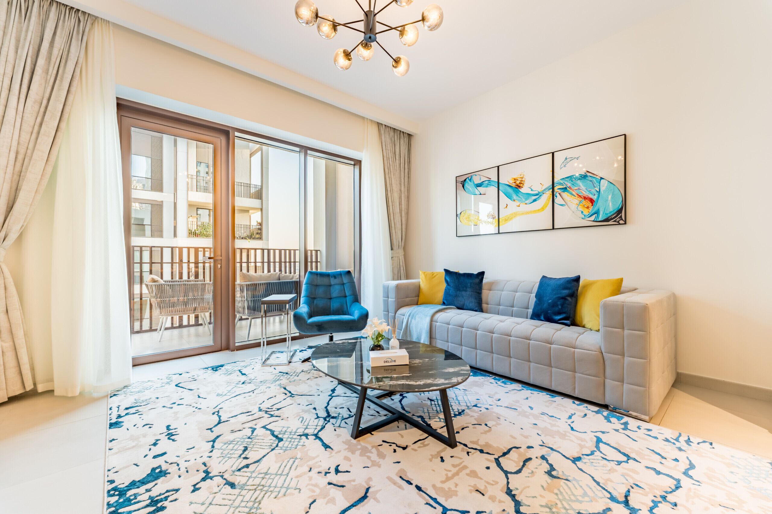 Una sala de estar moderna con un sofá gris, cojines azules y amarillos, una mesa de centro de vidrio y un cuadro abstracto grande.