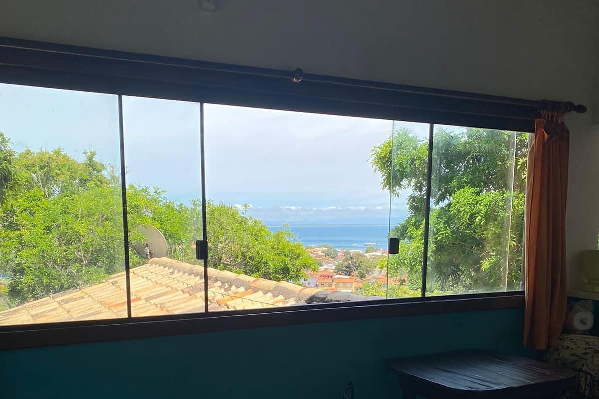 A room with a large window offering a view of greenery and the sea. There is a bed, a small table with a chair, and a lamp.