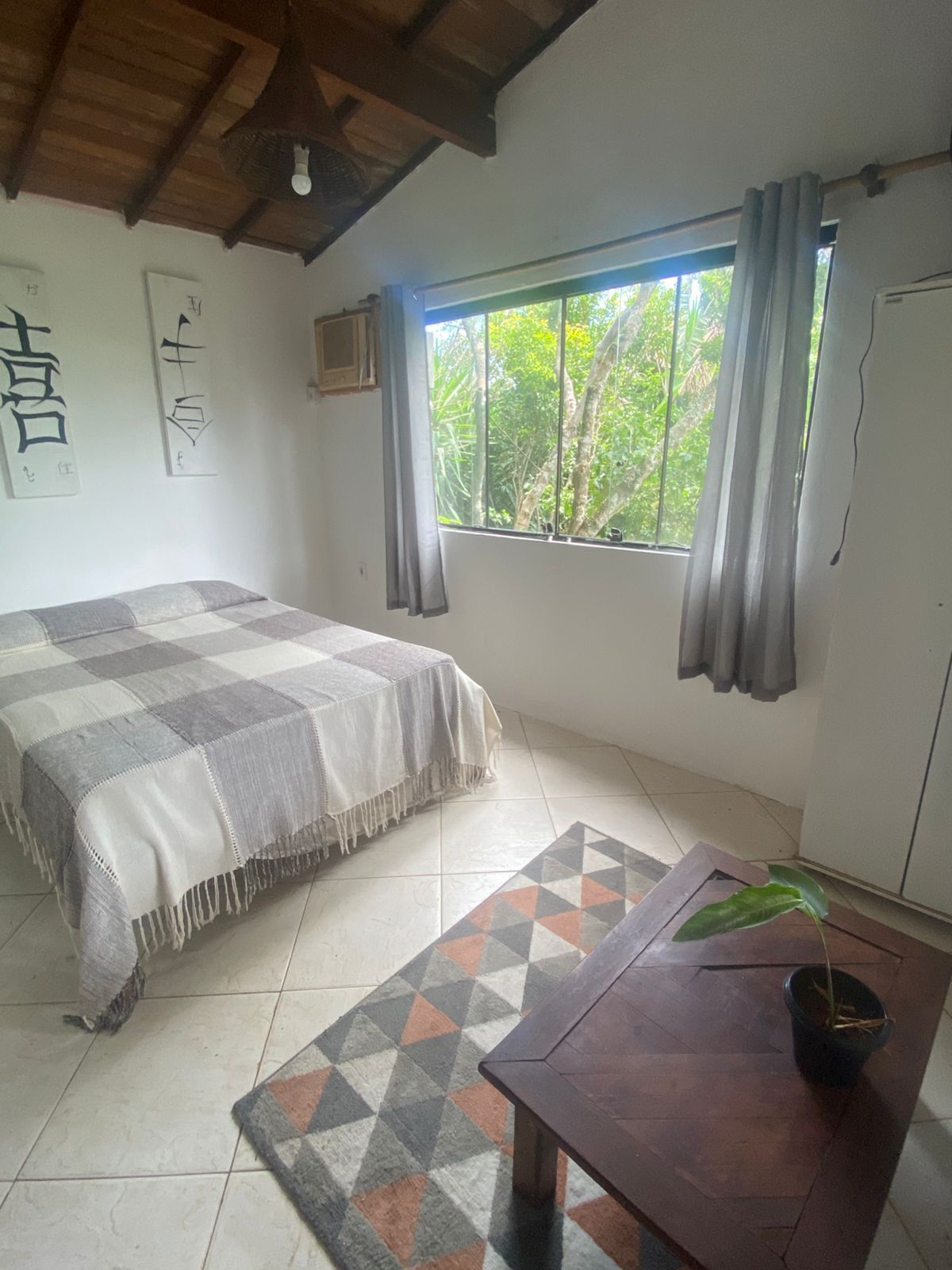 Un dormitorio con una cama, una mesa de madera, una planta en maceta, una ventana con cortinas y una pared con caligrafía.