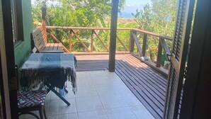 A covered outdoor area with a table and chairs, a wooden deck, and a view of greenery and a body of water. - Casamata (Búzios)