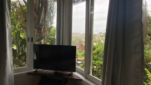 A bedroom with a bed, a TV on a stand, and a view of greenery and a distant landscape through large windows. - Casamata (Búzios)