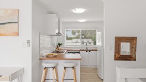 A modern kitchen with white cabinets, a wooden countertop, two bar stools, a window with blinds, and a hanging wooden frame. - Cerulean Getaway in Cowes (Cowes)