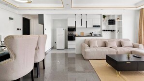 Een moderne woonkamer met een beige bank, een zwarte salontafel en een keuken met witte kasten en een koelkast.