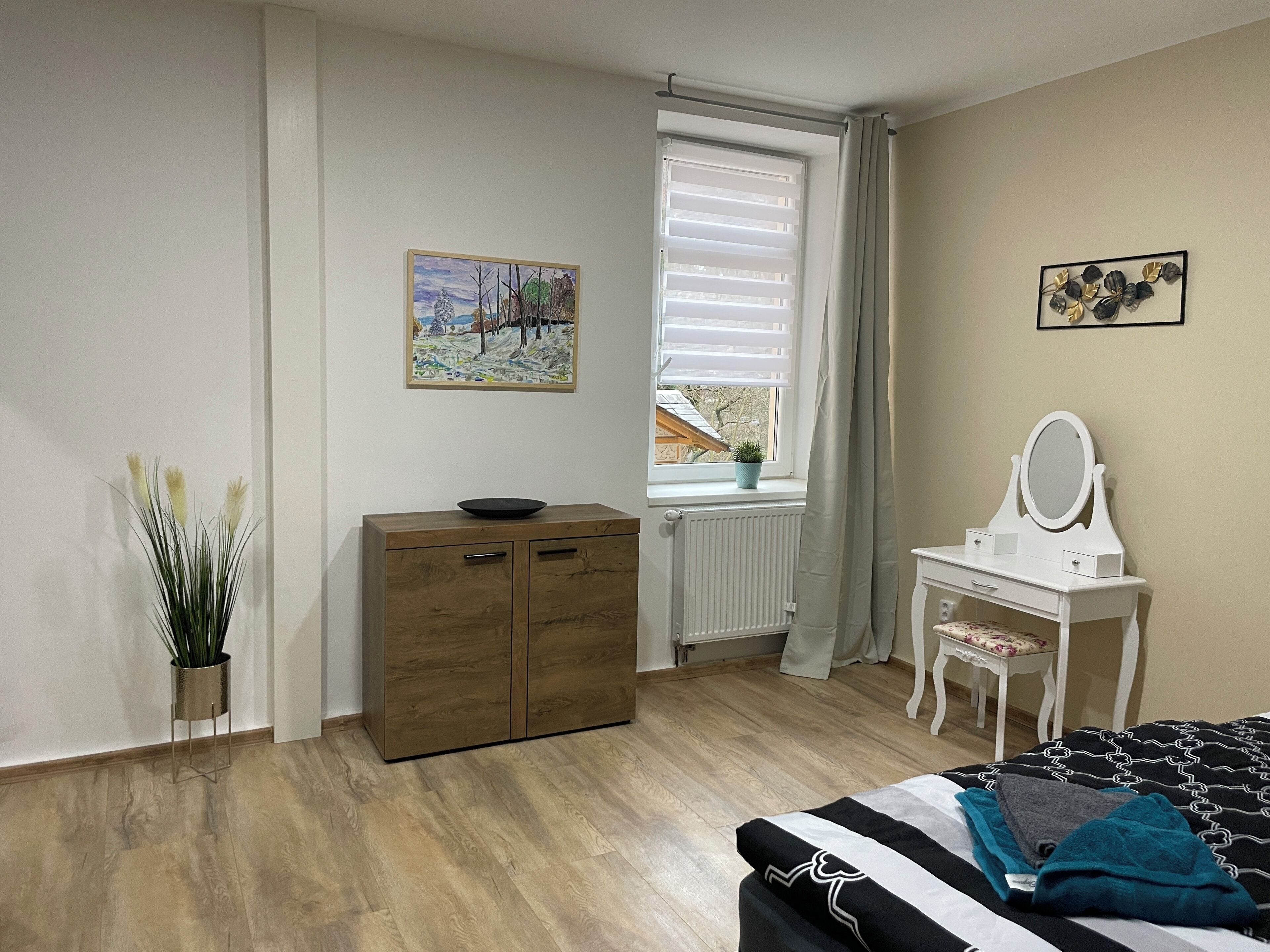 Een slaapkamer met een houten dressoir, een kaptafel met spiegel, een bed met zwart-wit gestreept beddegoed, een raam met zonwering en een schilderij aan de muur.