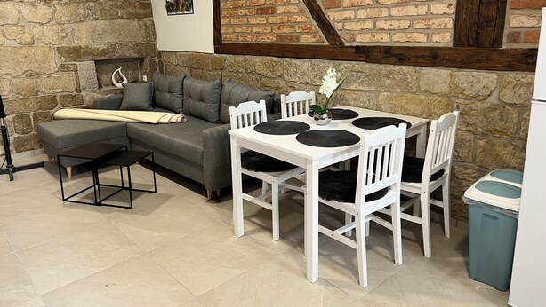 A cozy living area with a stone wall, a grey sofa, a white dining table with black placemats, and a small vase with flowers. - Apartments Badallee 4, Bad Schandau, apartment no. 8, 50m2 (Bad Schandau)