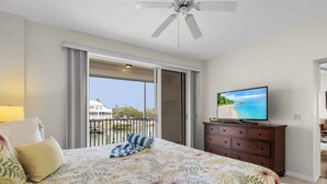 Un dormitorio con cama, televisión, cómoda y balcón con vista a la playa.