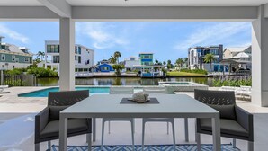 A modern outdoor patio with a pool, dining area, and a view of colorful buildings across a water body. - Brand New Luxury Build 265 Carolina Ave: Heated Pool, Hot Tub 6 Bedroom Home (Fort Myers Beach)