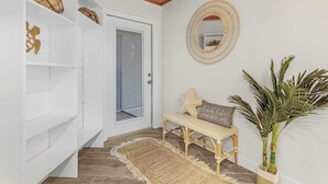 Un couloir avec une porte blanche, un miroir décoré de motifs tissés, un banc avec des coussins et une plante en pot.