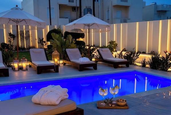 Un espace aménagé au bord de la piscine, avec des chaises longues, des parasols et une piscine dotée d’un éclairage bleu.