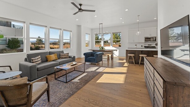 A spacious living room with a grey sofa, blue armchair, and a wooden coffee table. A kitchen area with white cabinets and a large window is visible in the background.