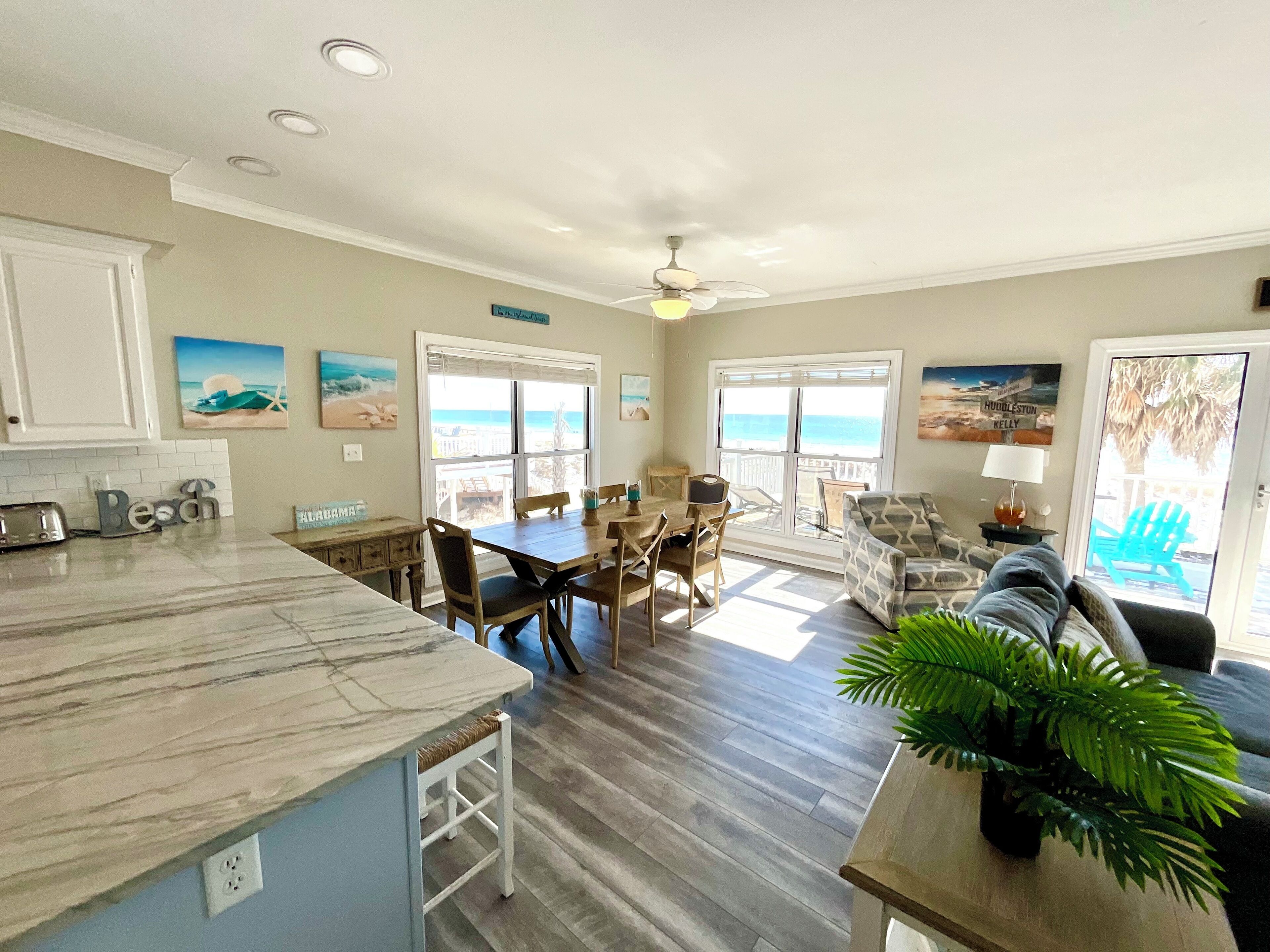 Una cocina amplia con encimera de mármol, una mesa de comedor de madera y sillas. Hay dos ventanas grandes con vistas a la playa.