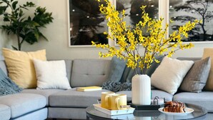 Ein modernes Wohnzimmer mit einem grauen Sofa, einem Glastisch und einer Vase mit gelben Blumen.