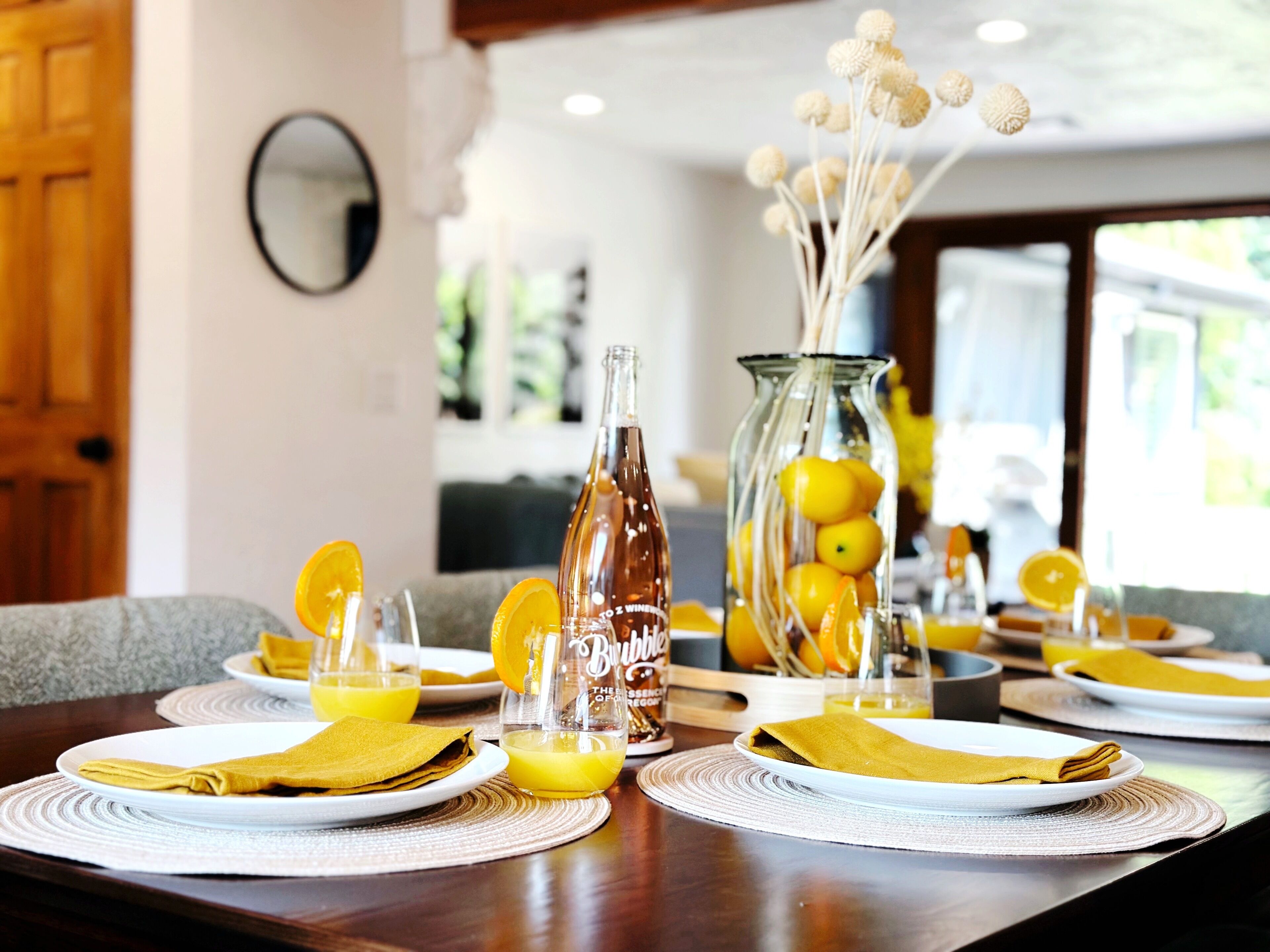 A dining table set with plates, napkins, and a bottle of Bubbly. There is a vase with lemons and decorative balls.