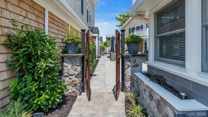 Un étroit sentier bordé de deux maisons aux murs recouverts de bardage en bois et agrémentées d’éléments en pierre, menant à une entrée fermée par un portail.