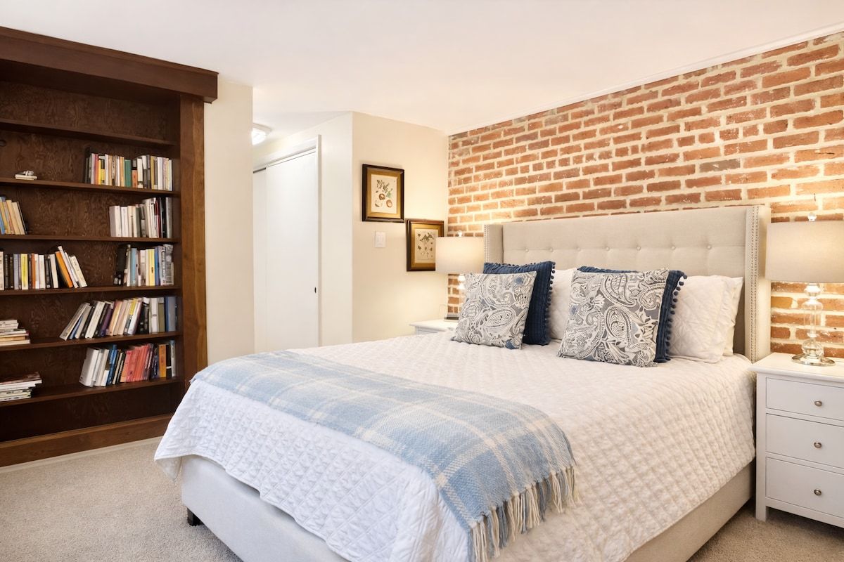 A bedroom with a brick wall, a large bed with patterned pillows, a bookshelf, and a nightstand with a lamp.