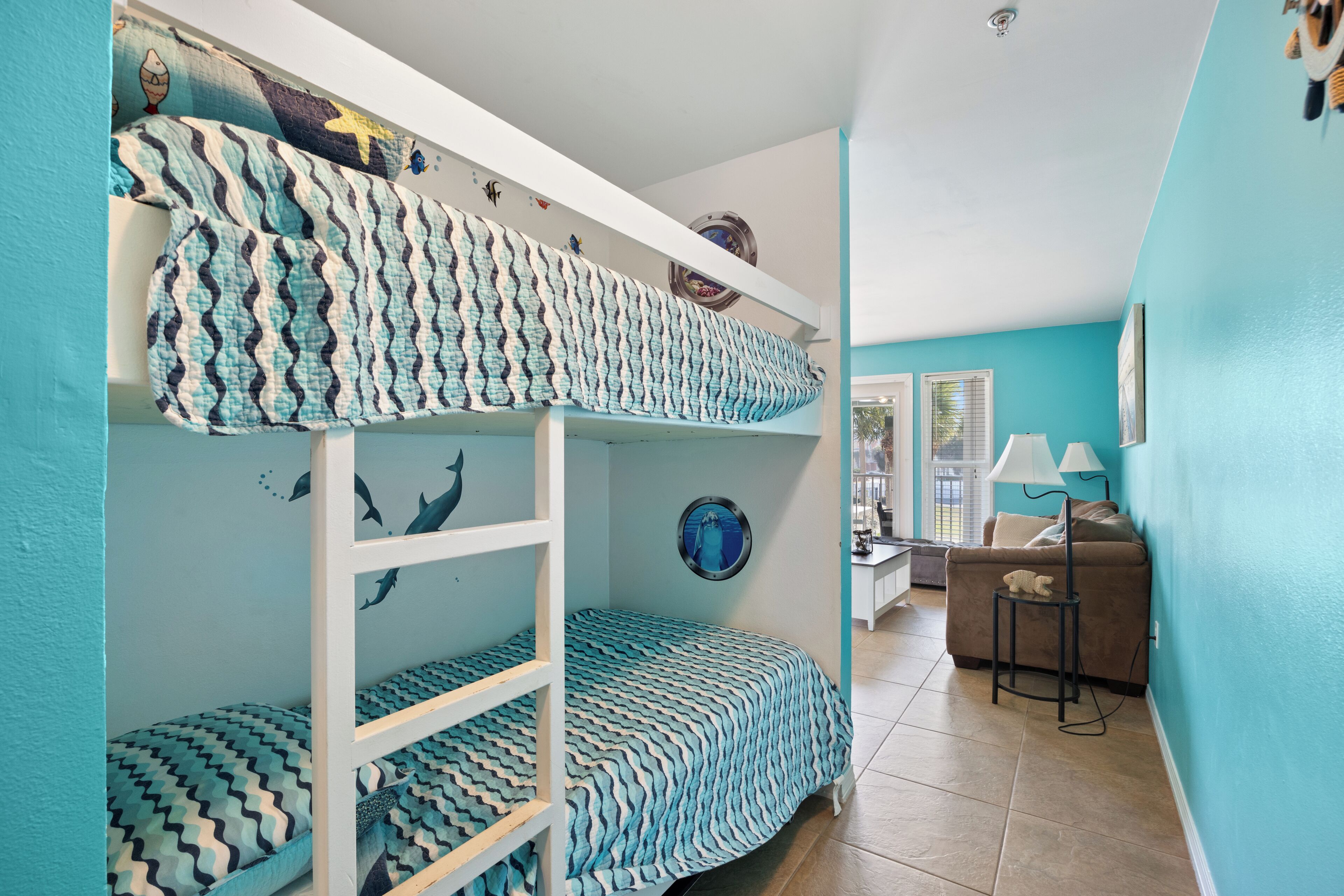 A bunk bed with patterned bedding, a desk with a lamp, and a window with blinds.