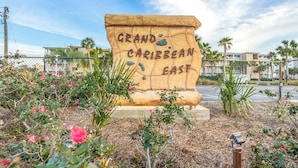 A large stone sign with the text "Grand Caribbean East" surrounded by flowering plants and a fence. - New Listing! Beach Vibe Is Only A Boardwalk Away From The Gulf! (Pensacola)
