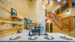 A wooden cabin interior with a central island dining table, built-in shelves, and a staircase. - Stylish Cabin Near Broken Bow Lake & Casino + Firewood Provided (Broken Bow)