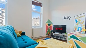 Una sala de estar con un sofá azul, una manta amarilla, un televisor mostrando 'Netflix' y un cartel de 'Los Simpson'.