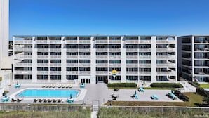 A multi-story hotel with a swimming pool, outdoor seating, and a clear blue sky.